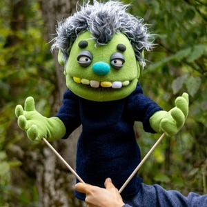 Könnte beinhalten: Eine grüne Monster-Handpuppe mit zerzaustem grauem Haar, einer blauen Nase sowie gelben und weißen Zähnen. Die Puppe trägt ein dunkelblaues Langarmshirt und hat grüne Hände mit jeweils vier Fingern. An den Händen sind zwei hölzerne Stäbe befestigt.
