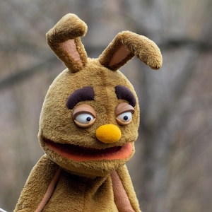 May include: A brown puppet with large, expressive eyes, a yellow nose, and a wide, orange mouth. The puppet has long, floppy ears and dark brown eyebrows. The puppet's fur is a soft, plush material.