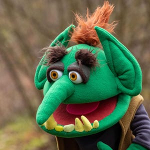 May include: A green puppet with large ears, a long nose, and a wide mouth. The puppet has yellow teeth, a red tongue, and brown eyebrows. It is wearing a brown vest and a black shirt. The puppet has orange hair.