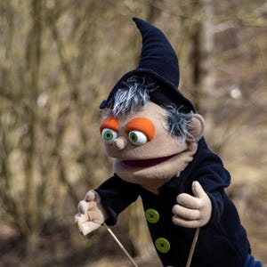 Könnte beinhalten: Eine Marionette mit einem beigen Gesicht, grünen Augen und orangefarbenem Lidschatten, die einen schwarzen Hut und Mantel mit grünen Knöpfen trägt. Die Marionette hat graues Haar und hält Fäden.