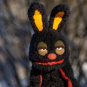 May include: A black bunny puppet with yellow inner ears, a red nose, and a red mouth. The puppet has brown eyes and eyebrows. The puppet has a red scarf. The background is blurred.