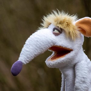 May include: A white puppet with a long nose and a purple tip. The puppet has a light brown hair and orange ears. The mouth is open, revealing an orange interior. A wooden stick is attached to the puppet.