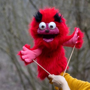 May include: A red puppet with large, expressive eyes, a pink mouth, and pink hands. The puppet has black tufts of hair and is made of fluffy material. It is held by wooden sticks, suggesting it is a hand puppet.