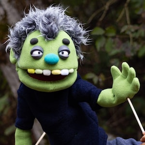 May include: A green puppet with messy grey hair, dark eyelids, and a blue nose. The puppet has a wide mouth with yellow and white teeth, wears a dark navy blue shirt, and is attached to a thin wooden control rod held by a hand at the bottom.