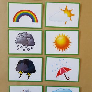 May include: A collection of weather-themed flashcards. Each card features a different weather condition, including clouds, a rainbow, rain, snow, and a sunny day. The cards have a green border and are arranged on a light brown surface.