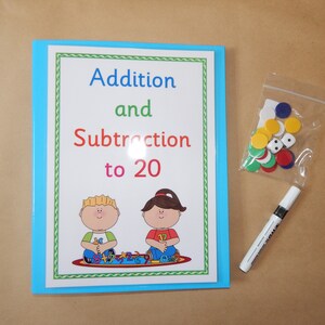 May include: A blue binder with a white cover featuring the text Addition and Subtraction to 20, illustrations of two children, and a green border. Included are a clear plastic bag of colourful plastic discs and two dice, plus a white dry-erase marker.