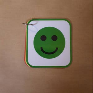 May include: A small stack of cards with a green smiley face on the front. The smiley face has two black eyes and a curved black mouth. The cards are held together by a metal ring.
