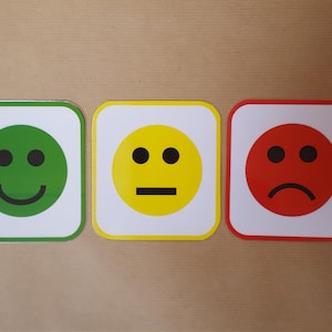 May include: Three square emoji signs with rounded corners. The first is green with a smiling face, the second is yellow with a neutral face, and the third is red with a sad face. The signs are on a light brown surface.