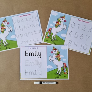May include: Three educational activity sheets featuring unicorn illustrations and tracing exercises. One sheet shows the alphabet, one shows numbers zero through ten, and one shows the name Emily. A black dry-erase marker is positioned below the sheets.