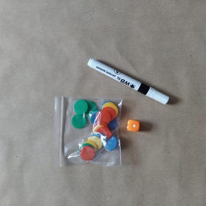 May include: A collection of colorful game pieces, including green, red, yellow, and blue circular tokens in a clear plastic bag, an orange die, and a white dry-erase marker with black accents.