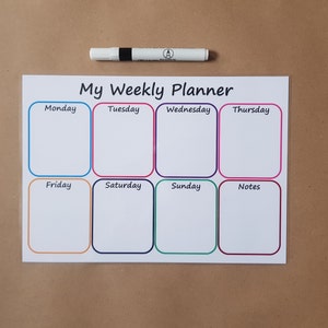 May include: A white weekly planner with the text "My Weekly Planner" and boxes for each day of the week, plus a notes section. A white marker with a black band is positioned above the planner. The boxes are outlined in various colours.