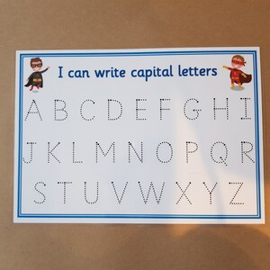 May include: A white educational chart with the text "I can write capital letters" and the alphabet in dotted lines. Superhero illustrations are in the corners. The chart is in a clear plastic sleeve.