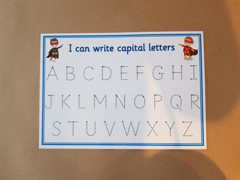 May include: A white educational chart with the text "I can write capital letters" and the alphabet in dotted lines. Superhero illustrations are in the corners. The chart is in a clear plastic sleeve.