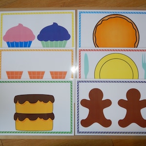 May include: A collection of illustrated food cards featuring a pink cupcake, a blue cupcake, a pancake, three orange waffle cones, a yellow plate with a knife and fork, a two-layer cake, and two gingerbread men.