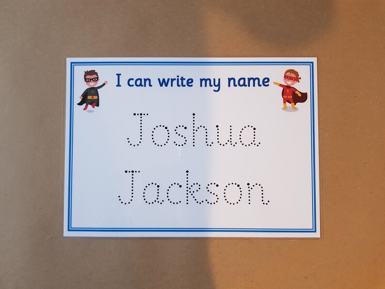 May include: A white educational learning board with a blue border and the text "I can write my name." The names "Joshua" and "Jackson" are printed in dotted lines for tracing. Superhero illustrations are on the top corners.
