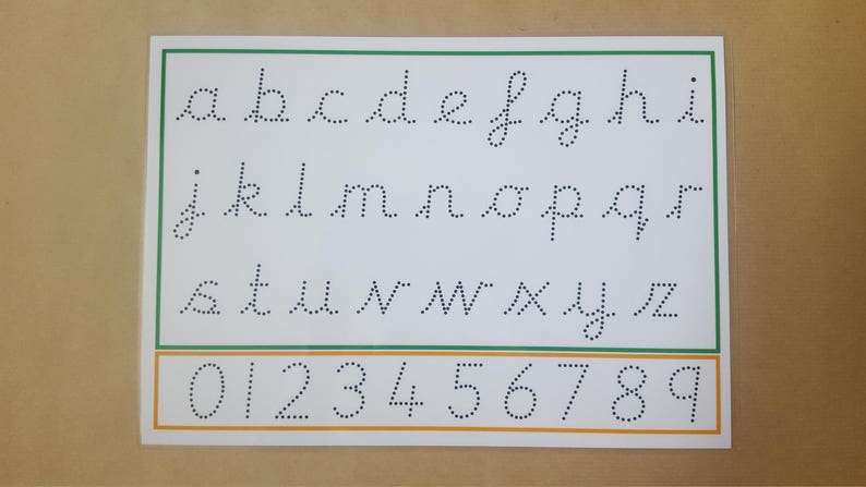 May include: A white educational chart with a green and orange border. The chart displays lowercase cursive letters a-z and numbers 0-9, all in dotted lines for tracing. The background is a light brown color.