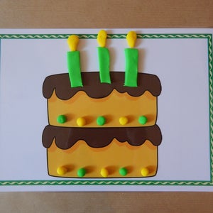 May include: A colorful birthday cake illustration with three candles. The two-tiered cake is yellow with brown frosting and decorated with green and yellow dots. The candles are green with yellow flames, set on a white background with a green border.