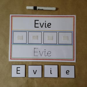 May include: A white educational board with the name "Evie" printed in black. The board has a red, blue, and green border, with four squares and a dotted line version of the name. Individual letter tiles spell "Evie" are below.