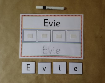 May include: A white educational board with the name "Evie" printed in black. The board has a red, blue, and green border, with four squares and a dotted line version of the name. Individual letter tiles spell "Evie" are below.