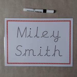 May include: A white card with the name "Miley Smith" written in dotted letters, framed by a red and white border. A white dry-erase marker with a black band and cap rests above the card.