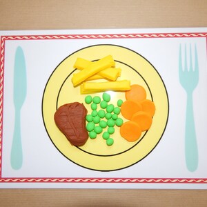 May include: A placemat featuring a yellow plate with a brown steak, green peas, orange carrot slices, and yellow fries. A light blue knife and fork are on either side of the plate. The placemat has a white border with a red zig-zag pattern.