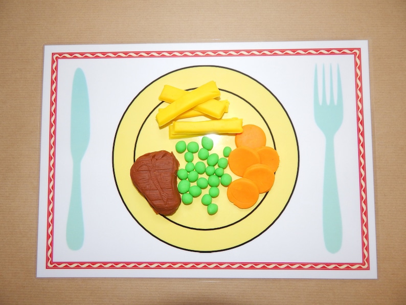 May include: A placemat featuring a yellow plate with a brown steak, green peas, orange carrot slices, and yellow fries. A light blue knife and fork are on either side of the plate. The placemat has a white border with a red zig-zag pattern.