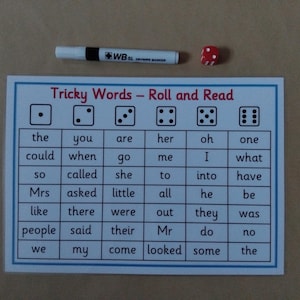 May include: A literacy game with a card displaying the text "Tricky Words - Roll and Read" and a grid of words. A red die and a white marker are placed above. A learning resource for children.