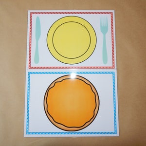 May include: Two illustrated placemats with a dining theme. The top placemat features a yellow plate, a light blue knife, and a light blue fork. The bottom placemat has an orange circle. Both placemats have decorative borders.