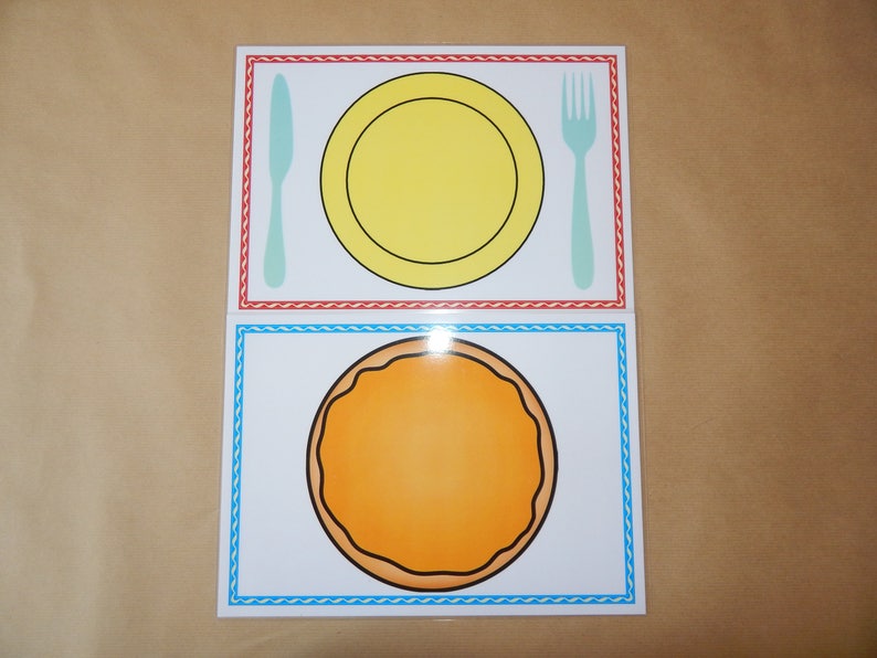 May include: Two illustrated placemats with a dining theme. The top placemat features a yellow plate, a light blue knife, and a light blue fork. The bottom placemat has an orange circle. Both placemats have decorative borders.