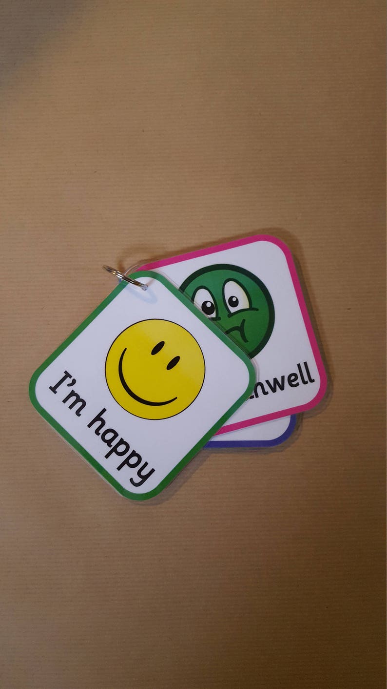 May include: Two square cards with rounded corners, attached by a metal ring. One card has a yellow smiley face and the text "I'm happy". The other card has a green face and the word "unwell". Both cards have white backgrounds.