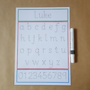 May include: A white educational worksheet with the name "Luke" at the top, featuring dotted lowercase letters and numbers for tracing. A white marker with a black band is placed next to the worksheet. The borders are green, red, and blue.