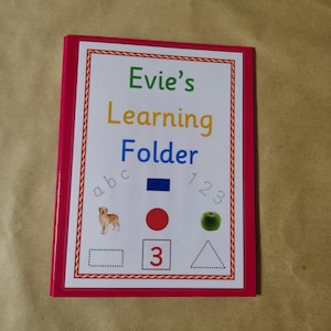 May include: A pink-edged learning folder labelled "Evie's Learning Folder" with educational illustrations. The cover displays the letters a, b, c, the numbers 1, 2, 3, a blue rectangle, a red circle, a green apple, and a dog.