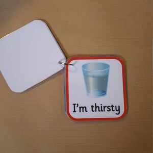 May include: A flashcard with a picture of a glass of water and the text "I'm thirsty". The card is attached to a blank white card with a metal ring. The cards have a red border.