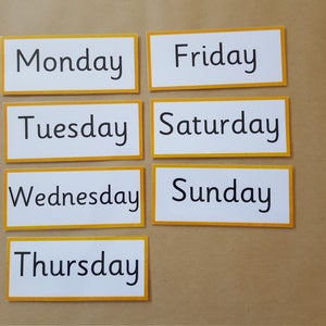 May include: Seven rectangular cards with the days of the week printed in black text on a white background. Each card has a yellow border. The cards are arranged on a brown surface.
