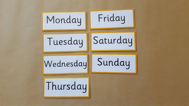 May include: Seven rectangular cards with the days of the week printed in black text on a white background. Each card has a yellow border. The cards are arranged on a brown surface.