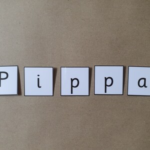 May include: Five white square tiles spelling the name "Pippa" in black capital letters and lowercase letters. The tiles are arranged horizontally on a light brown surface.