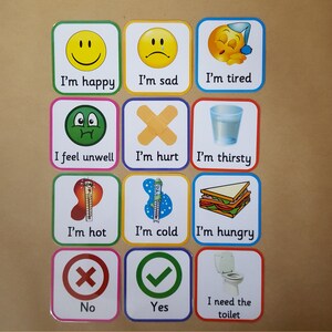 May include: A set of twelve square cards with various illustrations and text. The cards depict emotions and needs, including "I'm happy," "I'm sad," "I'm tired," "I feel unwell," "I'm hurt," "I'm thirsty," "I'm hot," "I'm cold," "I'm hungry," "No," "Yes," and "I need the toilet."