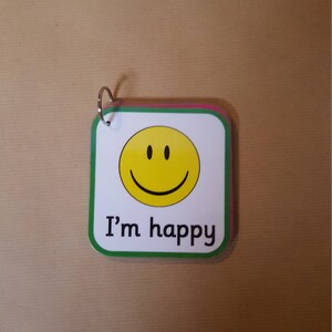 May include: A small, square card with a yellow smiley face and the text "I'm happy". The card has a white background with a green border and a silver ring at the top. The card is on a brown surface.