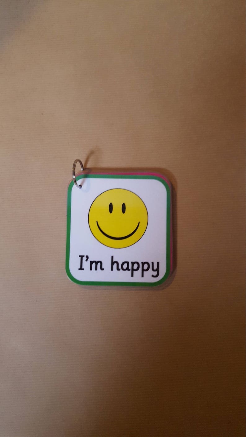 May include: A small, square card with a yellow smiley face and the text "I'm happy". The card has a white background with a green border and a silver ring at the top. The card is on a brown surface.