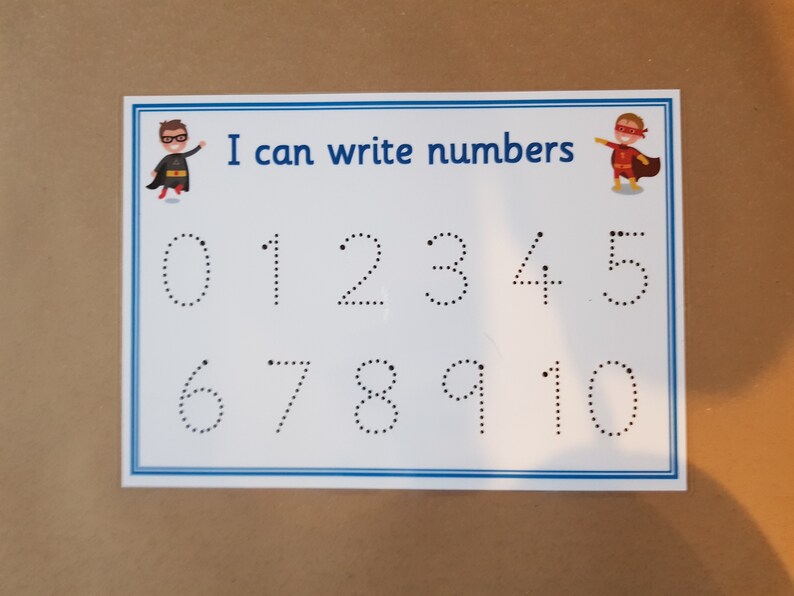 May include: A white educational card with a blue border, featuring the text "I can write numbers." Numbers 0-10 are displayed as dotted lines for tracing. Cartoon superheroes are in the corners.