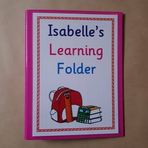 May include: A pink folder with a white cover. The cover has the text "Isabelle's Learning Folder" in blue and pink. A cartoon backpack, ruler, and books are also on the cover.