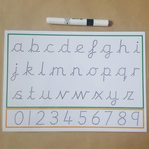 May include: A white handwriting practice sheet with dotted lowercase letters and numbers 0-9, outlined in green and orange. A white and black pen rests above the sheet, ideal for learning to write. The sheet is on a tan surface.
