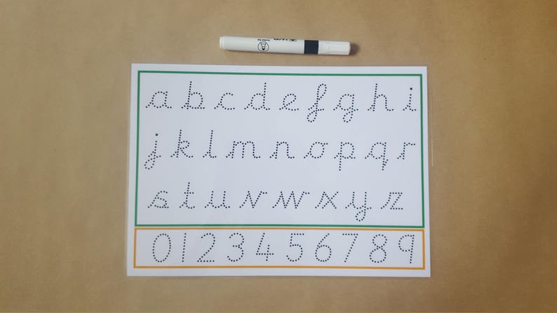May include: A white handwriting practice sheet with dotted lowercase letters and numbers 0-9, outlined in green and orange. A white and black pen rests above the sheet, ideal for learning to write. The sheet is on a tan surface.