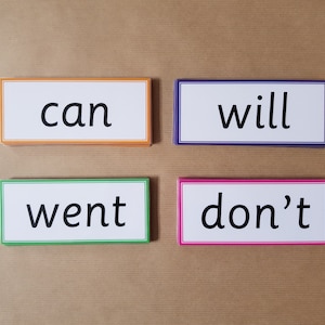 May include: Four rectangular flashcards with the words "can", "will", "went", and "don't" printed in black. Each card has a white background and a colored border: orange, purple, green, and pink. The cards are arranged on a brown surface.