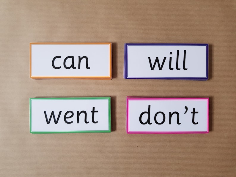 May include: Four rectangular flashcards with the words "can", "will", "went", and "don't" printed in black. Each card has a white background and a colored border: orange, purple, green, and pink. The cards are arranged on a brown surface.