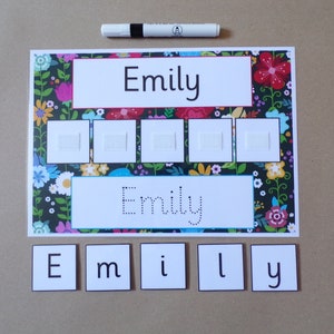 May include: A learning board with a floral border, displaying the name "Emily" in black. The board has spaces for attaching items and a dotted version of the name for tracing. Individual letter tiles spell "Emily" below. A black marker is at the top.