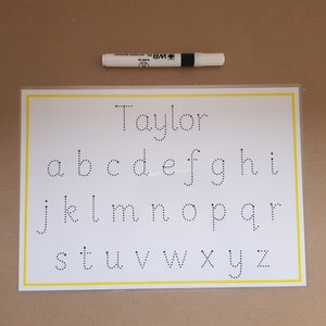 May include: A white educational worksheet featuring the lowercase alphabet in dotted letters, with the name "Taylor" above. A white dry-erase marker with black accents is positioned above the worksheet. The worksheet has a yellow border.