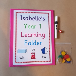 May include: A pink learning folder titled "Isabelle's Year 1 Learning Folder" with a red and white border. Includes a white marker, a red die, and a small bag of colourful counters. The folder has illustrations and text.