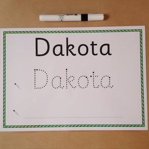 May include: A white sheet of paper with the name "Dakota" printed in large, black letters and dotted letters below for tracing. A white marker with a black cap rests above the paper. The paper has a green border.