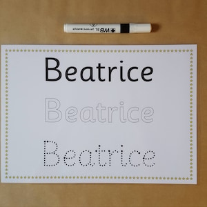 May include: A white sheet of paper with the name "Beatrice" printed in three different styles: solid, outlined, and dotted. A white WB dry erase marker is positioned above the paper. The paper has a gold star border.
